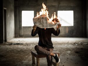 Muž sedící na židli drží v ruce hořící noviny