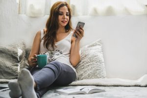 Žena drží telefon, sedí pohodlně, čte si zprávy a pije kafe