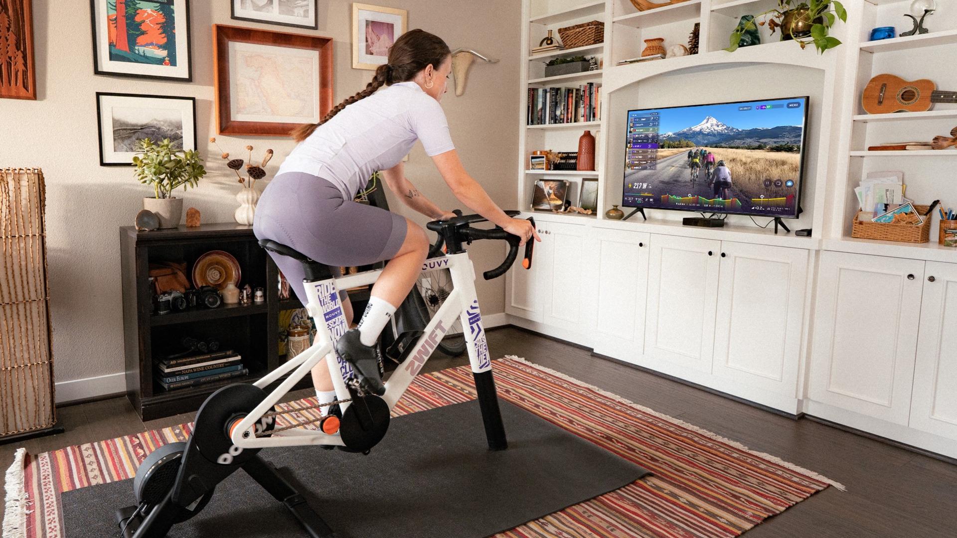 Cyklistka na bílém rámu Zwift Ride a trenažéru před televizí s trasou Rouvy ve světlém obýváku