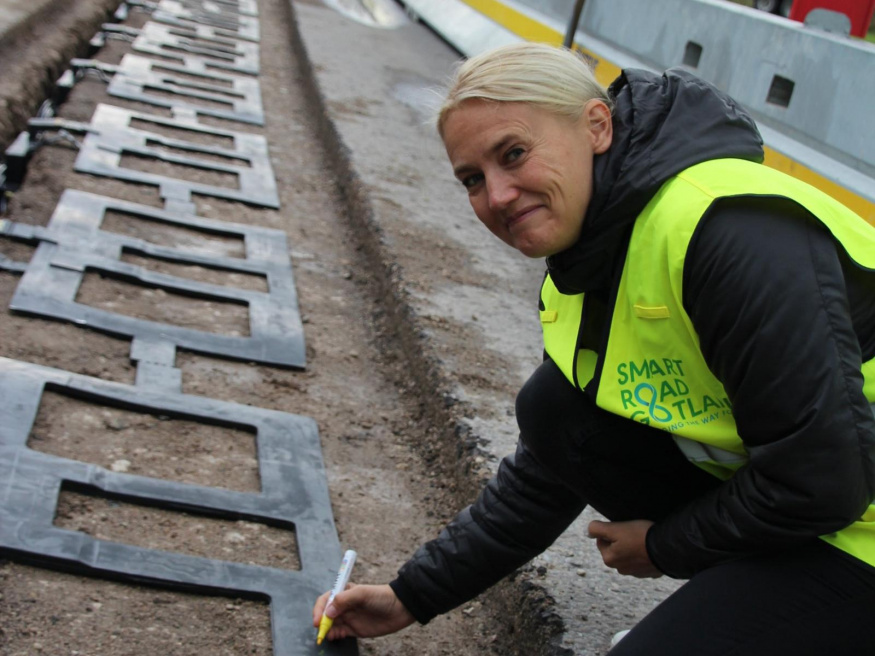 Cívky Electreon při stavbě silnice Smartroad Gotland Řada plochých nabíjecích cívek v silničním výkopu a žena v reflexní vestě u jedné z nich