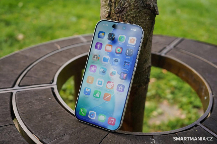 Displej základního iPhonu 17 Zapnutý iPhone 17 opřený o strom na kruhové dřevěné lavičce