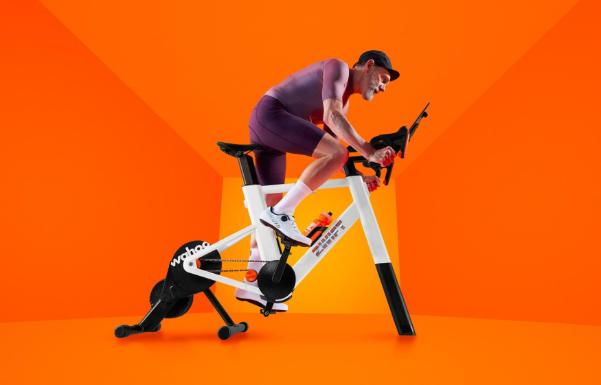 Sestava Zwift Ride s trenažérem Wahoo Cyklista na bílém rámu Zwift Ride s trenažérem Wahoo a tabletem před oranžovým pozadím