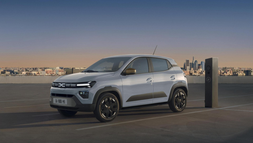 Modernizovaná Dacia Spring Světle modrá Dacia Spring na střešním parkovišti s městem v pozadí