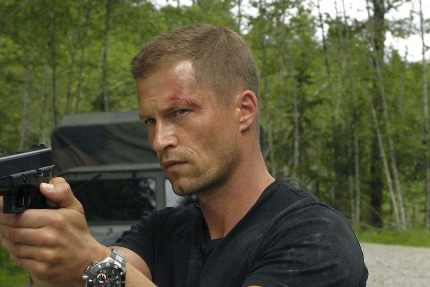 Jack Carver ve filmovém Far Cry Til Schweiger v černém tričku míří pistolí doleva, za ním jsou stromy a vozidlo