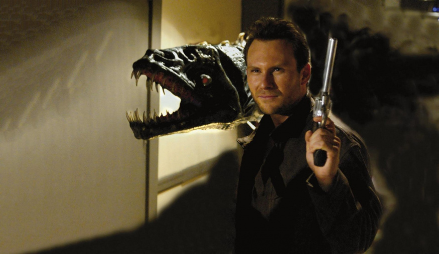 Edward Carnby ve filmu Alone in the Dark Christian Slater v tmavém kabátu drží pistoli vedle velké zvířecí lebky s otevřenou čelistí