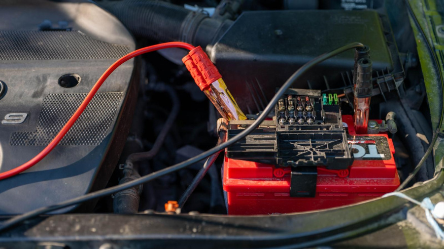 Autobaterie s připojenými startovacími kabely Červená autobaterie v motorovém prostoru s připojenými svorkami startovacích kabelů