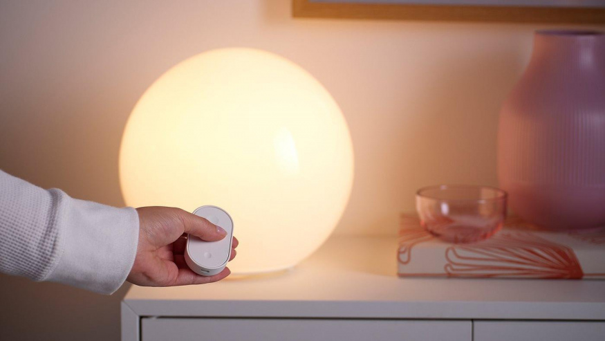 Sada IKEA KAJPLATS při ovládání světla Ruka s bílým ovladačem sady IKEA KAJPLATS před lampou svítící teplým bílým světlem
