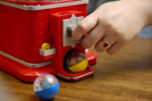 Výdej kapsle z LEGO automatu Ruka otáčí šedou klikou červeného LEGO automatu a žlutá kapsle leží ve výdejním otvoru