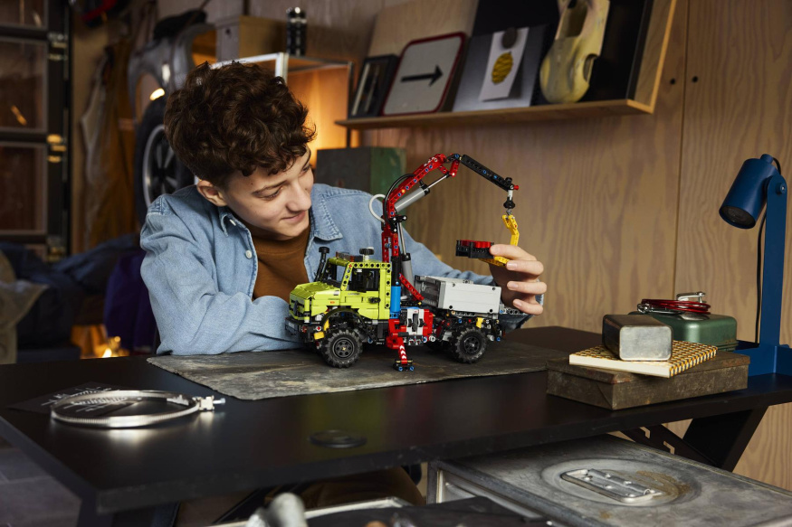 LEGO Unimog s pneumatickým jeřábem Mladík obsluhuje zelený LEGO Unimog s červeným kloubovým jeřábem, vysunutou podpěrou a paletou u háku