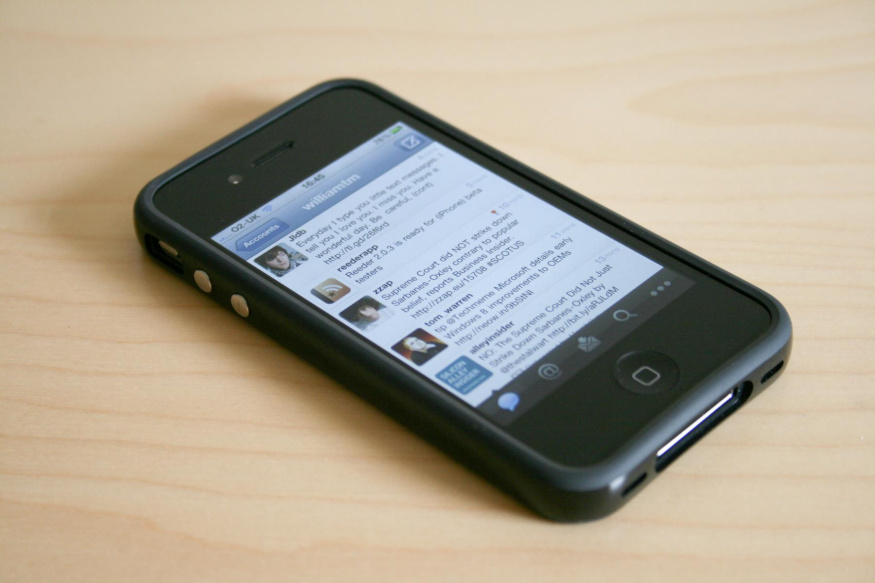 iPhone 4 s aplikací Twitter Černý iPhone 4 v ochranném rámečku na stole, s aplikací Twitter na displeji a viditelným tlačítkem Home
